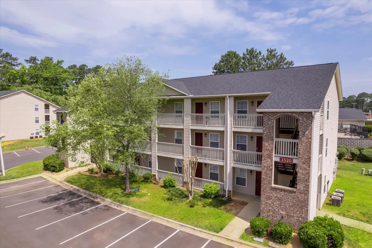 A bright and inviting exterior shot of the apartment building, surrounded by green landscaping and a convenient parking area. The image captures the building from a perspective that suggests easy access for commuters and business travelers alike.