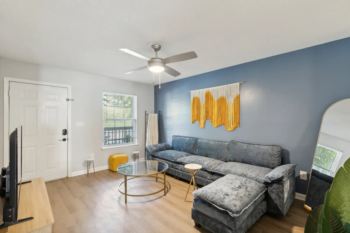 A cozy living area with a comfortable sofa, glass coffee table, modern wall art, and a wide window that bathes the room in natural light. The setup invites conversation, movie nights, or quiet moments of relaxation.