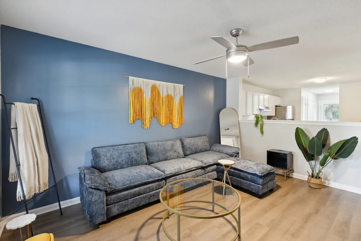 A bright, open living room featuring a contemporary blue sofa, modern glass coffee table, vibrant yellow wall hanging, and a lush green houseplant. Sunlight pours in through large windows, creating a cozy, inviting space perfect for relaxing or gathering with friends and family.