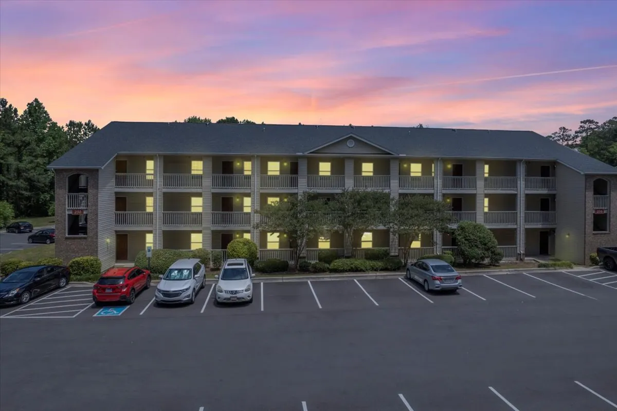 This exterior image features the apartment building with a well-kept parking lot, landscaped greenery, and a vibrant sunset sky overhead. The setting exudes a warm, inviting neighborhood feel and hints at easy accessibility for visitors.