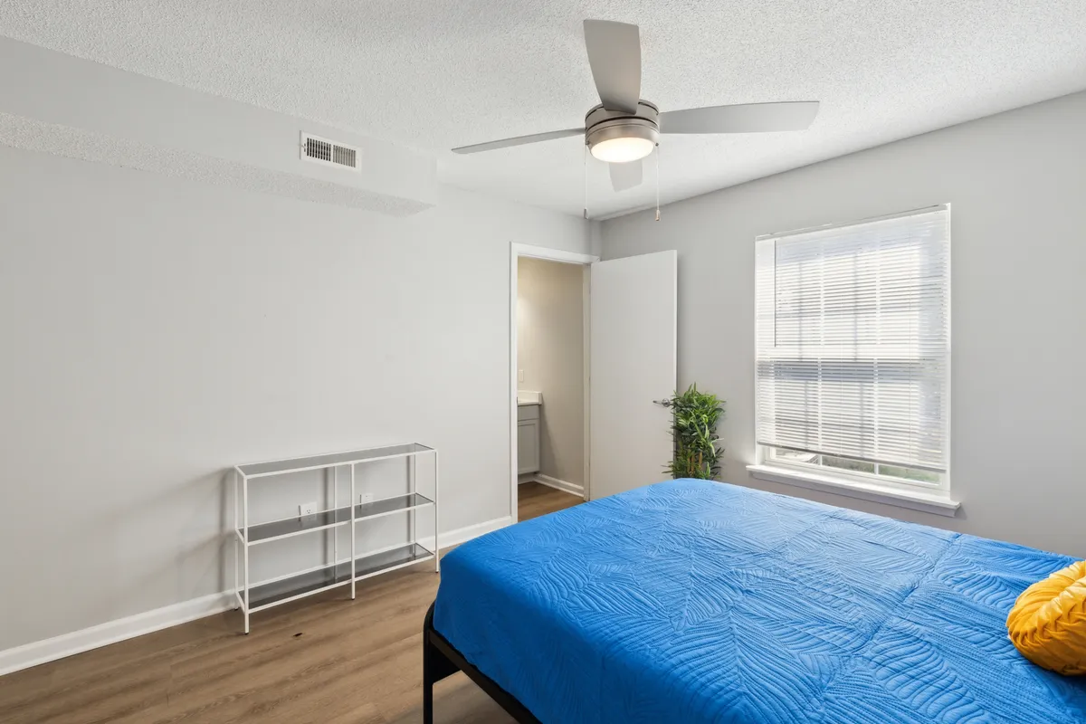 A tranquil bedroom featuring a blue bedspread, a white shelving unit, a green plant, and a window with blinds that lets in gentle natural light. The tidy, inviting setting offers a sense of relaxation and homey comfort.