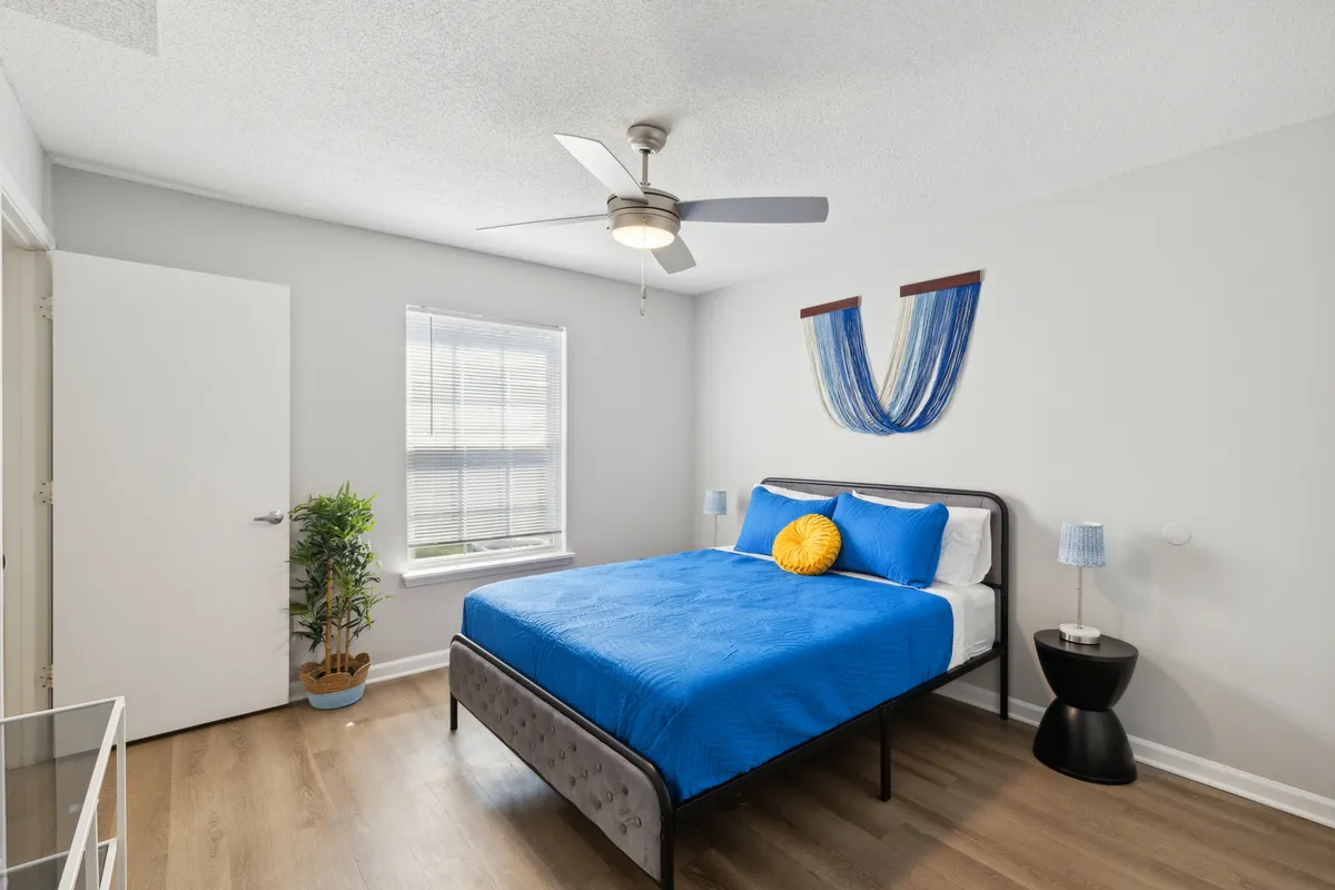 The image showcases a serene bedroom featuring a comfortable bed adorned with blue bedding and a cheerful yellow pillow, bathed in gentle natural light from a large window. Accented with modern decor and a ceiling fan overhead, the space feels calm, refreshing, and inviting.